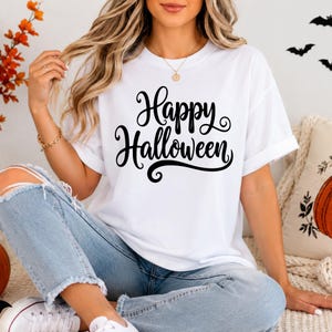 May include: A white short-sleeved t-shirt featuring the black text Happy Halloween in a cursive script font. The shirt has a crew neckline and rolled sleeves. The garment is paired with light blue denim jeans that have distressed knee detailing.