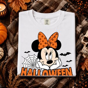 Puede incluir: Una camiseta blanca con un gráfico de un ratón de dibujos animados con un lazo naranja de lunares, una telaraña y varios murciélagos negros. La palabra HALLOWEEN está impresa en letras naranjas grandes en la parte inferior. Etiqueta Comfort Colors.