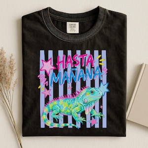 May include: A black t-shirt featuring a graphic of a green and blue iguana with pink spikes. Above the iguana, the text reads HASTA MAÑANA in pink and blue letters. The design includes light blue vertical stripes and pink star accents.