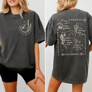 以下が含まれることがあります： チャコールグレーのTシャツ。左胸には三日月、星、ハーブの小さなグラフィックが描かれています。背面には、植物のイラスト、神秘的なシンボル、そして「Plant Roses & Lavender, for luck」「Keep Rosemary by your garden gate」というテキストが組み合わされた、大きく複雑なデザインがプリントされています。