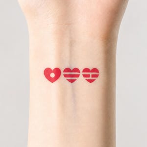 May include: Three red heart temporary tattoos on a wrist. The first heart has a dot in the center, the second has horizontal lines, and the third has a horizontal and vertical line.