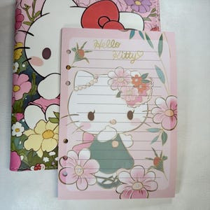 Puede incluir: Un juego de cuaderno y bloc de notas Hello Kitty rosa. La cubierta del cuaderno presenta un diseño floral con Hello Kitty. El bloc de notas tiene una ilustración de Hello Kitty rodeada de flores y las palabras "Hello Kitty".