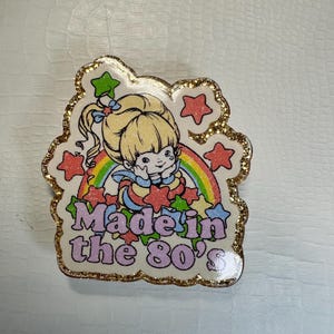 Op de afbeelding: Speld met een cartoonmeisje, regenboog en sterren. De tekst "Made in the 80's" is in roze en blauw. De speld heeft een gouden glitterrand.