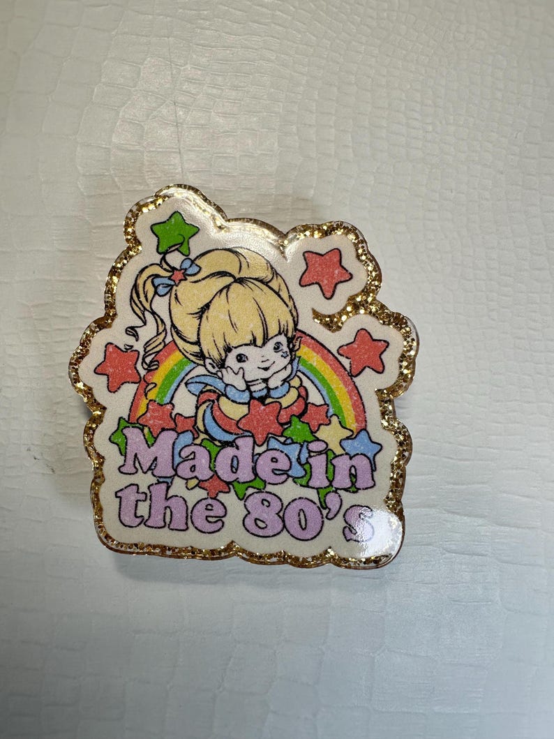 Op de afbeelding: Speld met een cartoonmeisje, regenboog en sterren. De tekst "Made in the 80's" is in roze en blauw. De speld heeft een gouden glitterrand.