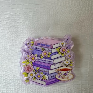 Op de afbeelding: Decoratieve afbeelding van een stapel boeken in paarse en roze tinten, versierd met bloemen. Bevat een kop en schotel. De boekruggen zijn beschreven, omringd door bloemmotieven.