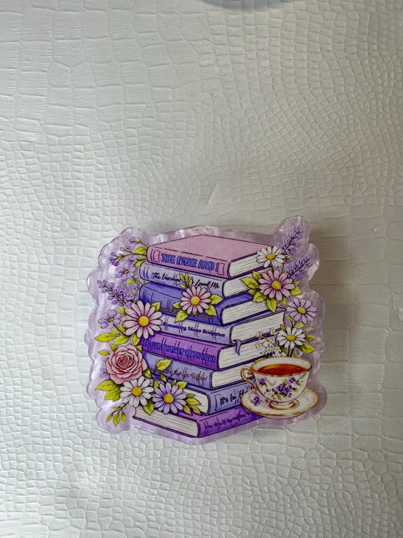 Op de afbeelding: Decoratieve afbeelding van een stapel boeken in paarse en roze tinten, versierd met bloemen. Bevat een kop en schotel. De boekruggen zijn beschreven, omringd door bloemmotieven.