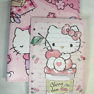 May include: Pink Hello Kitty themed notebook and binder set. The binder features a picnic scene and Hello Kitty. The notebook has a Hello Kitty graphic with cherries and the words "Cherry Jam".