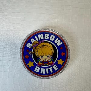 Op de afbeelding: Ronde container met een zilveren rand en een blauw midden. Het bevat de tekst "RAINBOW BRITE" en een cartoonafbeelding van een blond meisje. Het ontwerp bevat sterren en een regenboog, met een glitterrand.