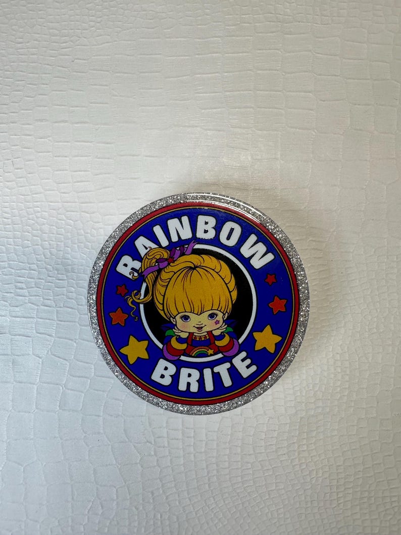 Op de afbeelding: Ronde container met een zilveren rand en een blauw midden. Het bevat de tekst "RAINBOW BRITE" en een cartoonafbeelding van een blond meisje. Het ontwerp bevat sterren en een regenboog, met een glitterrand.