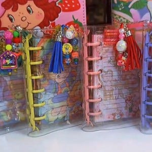 May include: Four clear plastic disc-bound notebooks featuring colorful spines in purple, yellow, pink, and light purple. Each notebook contains lined paper with cartoon character illustrations and is decorated with hanging charms, beads, and tassels.