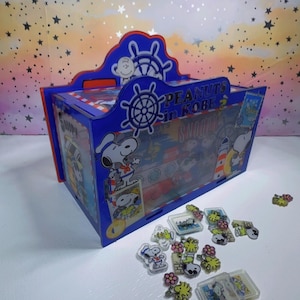 May include: A blue rectangular storage box featuring Peanuts in Kobe and Snoopy graphics with a ship wheel design. The box is decorated with illustrations of Snoopy in a sailor outfit and a lighthouse. Small clear acrylic charms are scattered nearby.