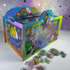 May include: A rectangular acrylic storage box featuring Rainbow Brite character graphics, rainbows, and stars, with the text Rainbow Brite printed on the sides. Several small, clear acrylic charms with printed character designs are scattered in front.