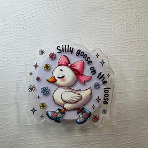 Op de afbeelding: Een heldere acryl sticker met een cartoon gans met een roze strik, die bloemenlaarzen draagt. De tekst "Silly goose on the loose" staat op de sticker, omringd door kleurrijke bloemen en sterren.