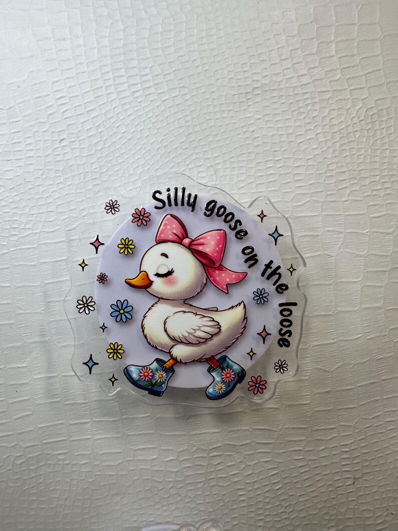 Op de afbeelding: Een heldere acryl sticker met een cartoon gans met een roze strik, die bloemenlaarzen draagt. De tekst "Silly goose on the loose" staat op de sticker, omringd door kleurrijke bloemen en sterren.