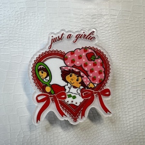 Op de afbeelding: Transparante acrylspeld met een cartoonmeisje met een aardbeienhoed en de tekst "just a girlie". De hartvormige speld heeft rode accenten, een rode strik en een spiegel. Het meisje houdt een spiegel vast en draagt een witte jurk.