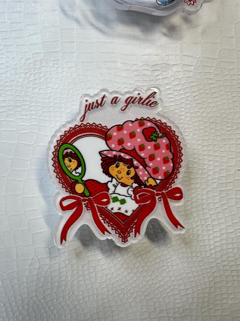Op de afbeelding: Transparante acrylspeld met een cartoonmeisje met een aardbeienhoed en de tekst "just a girlie". De hartvormige speld heeft rode accenten, een rode strik en een spiegel. Het meisje houdt een spiegel vast en draagt een witte jurk.