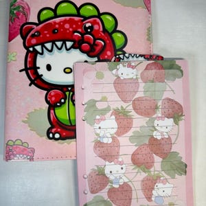 Puede incluir: Carpeta y cuaderno rosas con Hello Kitty. La carpeta tiene un diseño de disfraz de dinosaurio rojo. El cuaderno tiene un fondo rosa con ilustraciones de fresas y Hello Kitty. El cuaderno tiene páginas rayadas.