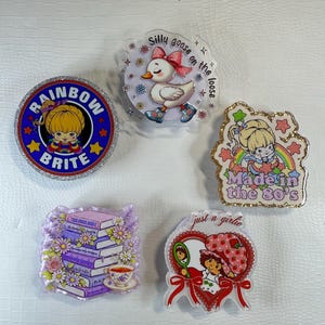 May include: A collection of five glittery acrylic pins featuring cartoon characters and phrases. Designs include a duck with a bow, a girl with the text "Rainbow Brite", and a girl with the text "Made in the 80's".