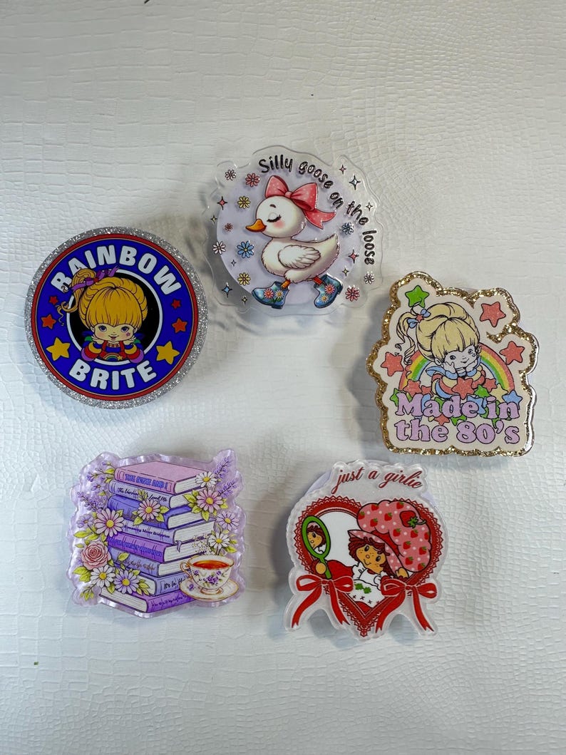 Op de afbeelding: Vijf acrylspelden met glitterranden, elk met een ander ontwerp. De spelden tonen stripfiguren en zinnen, waaronder een eend, een meisje met de tekst "Rainbow Brite" en "Made in the 80's".
