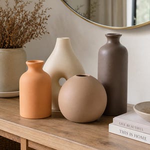 Pode incluir: Uma coleção de quatro vasos de cerâmica em várias formas e tons terrosos. O conjunto inclui um vaso laranja alto em forma de garrafa, um vaso creme com uma abertura central, um vaso esférico bege e um vaso cilíndrico castanho escuro.