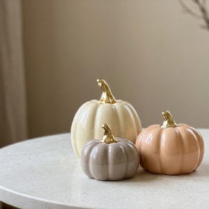 May include: A set of three ceramic pumpkins with glossy finishes in cream, soft peach, and muted taupe. Each pumpkin features a smooth, rounded body with vertical ridges and a reflective, metallic gold-coloured stem at the top centre.