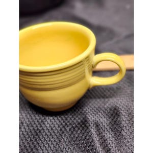May include: A yellow ceramic teacup with a rounded handle. The cup has a smooth, glossy finish and is decorated with horizontal, dark lines around the middle. The cup is set against a textured, grey background.