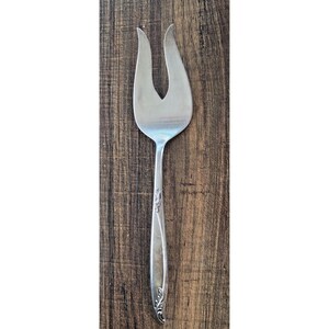 May include: A silver serving fork with a two-pronged design. The fork has a long, slender handle with decorative floral detailing. The background is a dark wood surface.