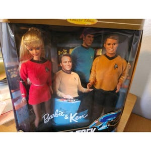 Puede incluir: Un set de edición coleccionista de Barbie y Ken en caja con personajes de Star Trek. El set incluye una muñeca Barbie con uniforme rojo y muñecos Ken con uniformes dorados y azules. La caja tiene las palabras "Barbie & Ken" y "Star Trek".