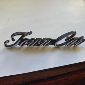 Puede incluir: Un emblema cromado "Town Car", probablemente para un vehículo. La escritura es en cursiva, con un fondo oscuro y una superficie brillante y reflectante. El emblema está diseñado para ser montado en un coche.