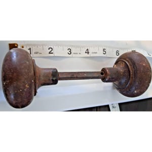 May include: A pair of dark brown, weathered metal door knobs connected by a square spindle. A white measuring tape is positioned behind the hardware, showing the total length of the assembly is approximately 6.5 inches from end to end.