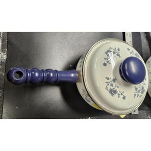 May include: A white enamel saucepan with a blue floral pattern and a matching blue handle and lid knob. The handle is a solid, rounded design. The pot has a silver rim.