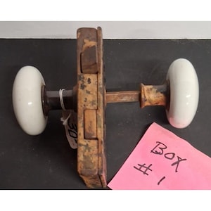 May include: A vintage door hardware set featuring two smooth white porcelain knobs attached to a central rectangular metal latch mechanism with a weathered, rusted finish. A small white tag is attached to one side, and a pink note reads BOX #1.