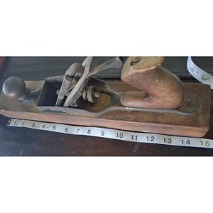 May include: A vintage wood-body hand plane with a metal blade assembly and a wooden handle. A white measuring tape is positioned along the base, showing the tool has a length of approximately 15 inches. The plane features a dark brown wooden body.