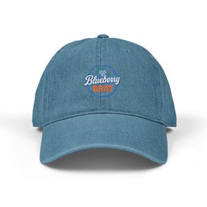 May include: A blue baseball cap featuring the text "Blueberry Brat" in white script and orange block letters. The text is within a blue circle with a blueberry design. The cap is made of a soft material and has a curved peak.