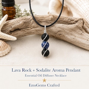 May include: A necklace featuring a black cord and a pendant composed of two black porous lava rock beads and one central blue sodalite bead, all wrapped in silver-toned wire. The text reads: Lava Rock + Sodalite Aroma Pendant Essential Oil Diffuser Necklace EmsGems Crafted.