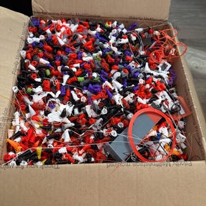May include: A cardboard box filled with a large collection of small, multi-colored 3D printed plastic components in red, white, black, purple, and orange, featuring thin filament strands and a single larger grey rectangular piece with a red ring.