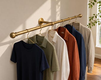 May include: A wall-mounted brass clothing rack with a cylindrical rod and rounded end caps, holding five metal hangers. The hangers support a navy t-shirt, an olive green polo shirt, a white button-down shirt, a rust-colored jacket, and a navy jacket.