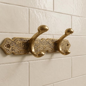May include: A brass-toned metal wall hook rack featuring two curved hooks attached to a rectangular backplate with an intricate embossed floral pattern and two visible screw holes for mounting on a white tiled wall surface.