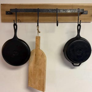 Könnte beinhalten: Ein hölzernes Küchenregal mit vier schwarzen Metallhaken, an denen zwei schwarze gusseiserne Bratpfannen und ein Holzschneidebrett hängen. Das Regal ist an einer hellen Wand montiert und zeigt eine rustikale Küchenästhetik.
