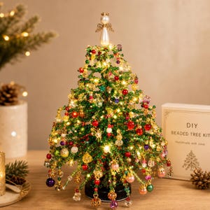 Op de afbeelding: Een decoratieve kerstboom gemaakt van groene kralen, versierd met veelkleurige kraalornamenten en geïntegreerde warmwitte lichtjes. Een witte parel engel figuur zit aan de top. Een doos met het label DIY BEADED TREE KIT is zichtbaar.