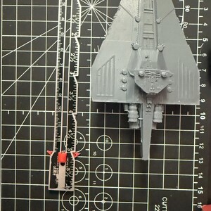 May include: A grey plastic spaceship model, approximately 13 cm long, sits on a black cutting mat. A ruler is placed beside the model for scale. The mat features a grid pattern and circular markings.