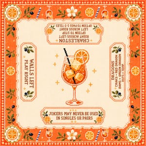 Op de afbeelding: Een vierkant borduurwerk met een oranje cocktail in het midden, inclusief ijsblokjes en een schijfje sinaasappel. De rand bevat sinaasappelpartjes, bloemen en tekstinstructies voor een spel, waaronder de regels voor Charleston, Dragons en Jokers.