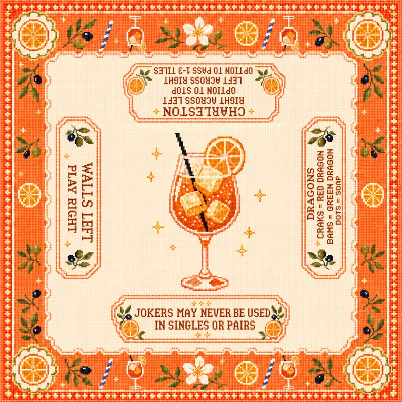 Op de afbeelding: Een vierkant borduurwerk met een oranje cocktail in het midden, inclusief ijsblokjes en een schijfje sinaasappel. De rand bevat sinaasappelpartjes, bloemen en tekstinstructies voor een spel, waaronder de regels voor Charleston, Dragons en Jokers.