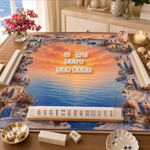K&ouml;nnte beinhalten: Ein Mahjong-Set mit einem mediterranen Landschaftsdesign. Das Spielbrett zeigt einen Sonnenuntergang &uuml;ber dem Meer, mit Geb&auml;uden und Blumen. Das Set enth&auml;lt Spielsteine, Leisten und eine kleine Schale mit Spielsteinen.