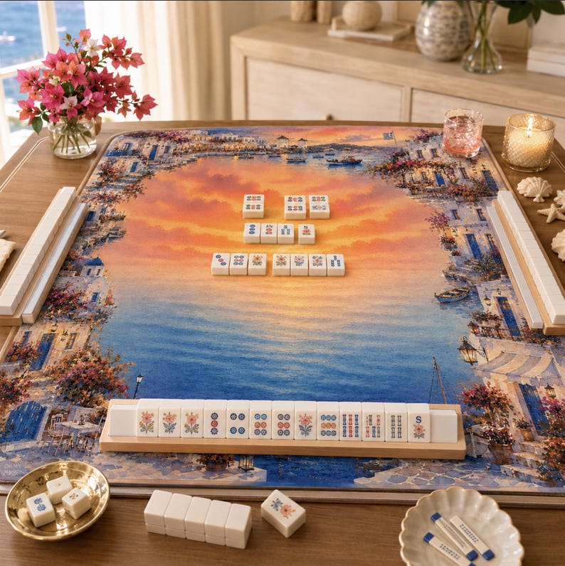 K&ouml;nnte beinhalten: Ein Mahjong-Set mit einem mediterranen Landschaftsdesign. Das Spielbrett zeigt einen Sonnenuntergang &uuml;ber dem Meer, mit Geb&auml;uden und Blumen. Das Set enth&auml;lt Spielsteine, Leisten und eine kleine Schale mit Spielsteinen.