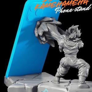 Peut inclure: Un support de téléphone gris représentant Goku de DBZ en position Kamehameha, tenant un téléphone bleu. Le support est sur une base grise texturée avec le texte "DBZ GOKU KAMEHAMEHA Phone stand" en orange et blanc.