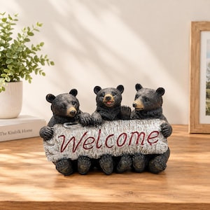 May include: A decorative figurine featuring three black bear cubs sitting behind a textured white birch log. The log has the word Welcome embossed in red script lettering. The bears have dark fur, tan muzzles, and are positioned in a row.