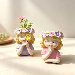 May include: Two resin planters shaped like children with blonde hair and floral crowns. The left planter features a child holding a yellow flower, while the right planter shows a child with a finger to their lips in a shushing gesture.