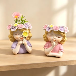 Può includere: Due vasi in resina a forma di bambini con capelli biondi e corone di fiori. Una figura indossa una maglietta viola e tiene una margherita gialla davanti a un occhio. L'altra figura indossa una maglietta rosa e porta un dito alle labbra in silenzio.