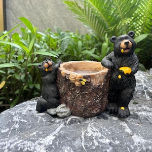 以下が含まれることがあります： 木株のプランターと黒いクマの置物セット。クマがプランターに手を伸ばし、もう一匹はハニカムを持っています。プランターは、蜂のアクセントが付いた、質感のある茶色の仕上がりです。